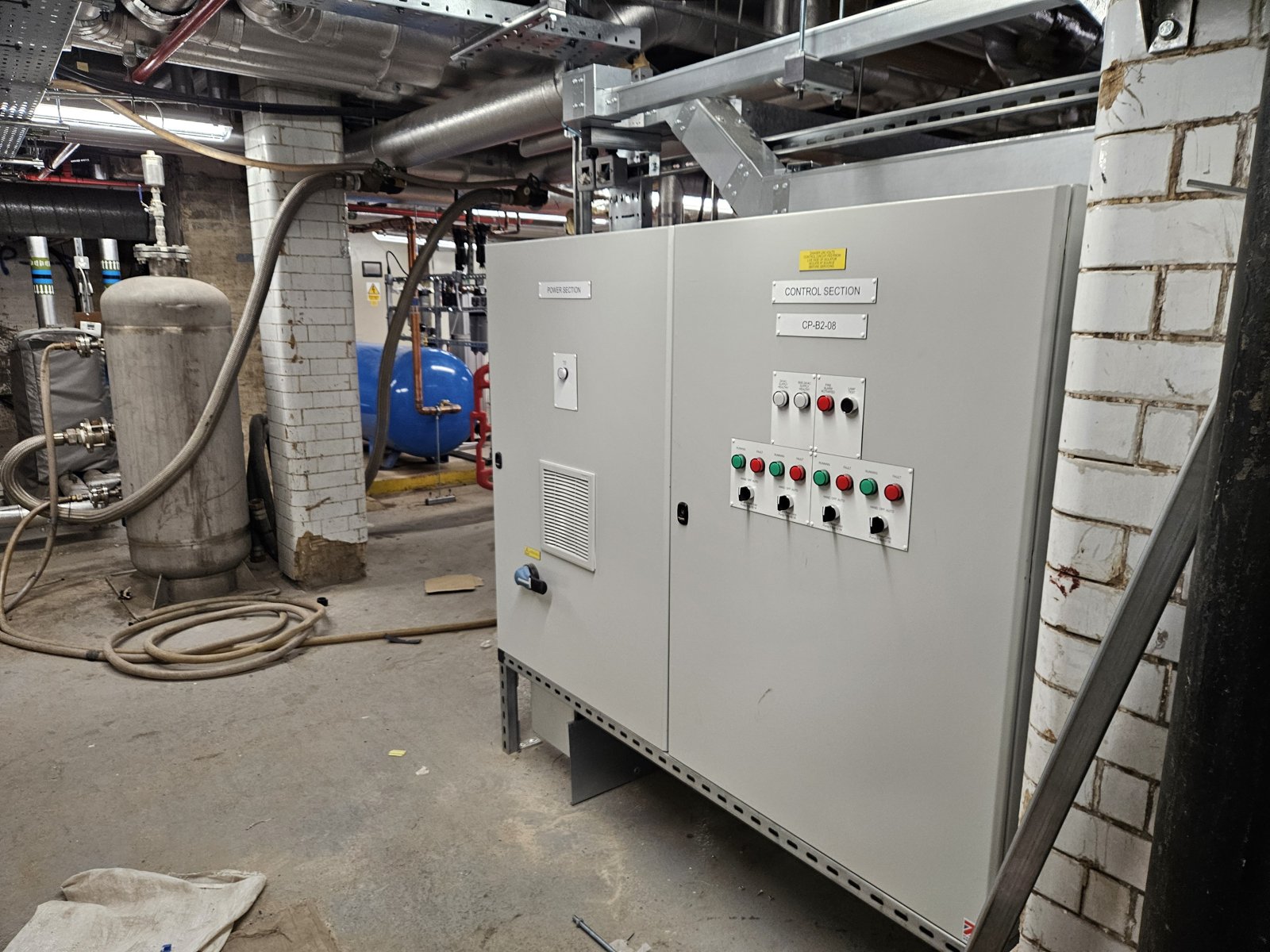 A two-door floor-standing BMS control panel installed in a plant room among pipework and plant